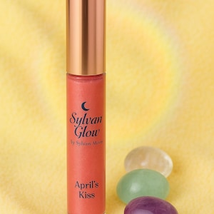 May include: A lip gloss tube with a copper-colored cap and a coral-colored body. The text "Sylvan Glow by Sylvan Moon" and "April's Kiss" are printed on the tube. Three oval-shaped gemstones in clear, green, and purple are next to the tube.