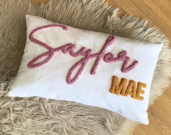 May include: White pillow with the name "Sayler Mae" embroidered in pink and yellow yarn.