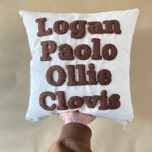 May include: White pillow with brown embroidered lettering that reads "Logan Paolo Ollie Clovis".