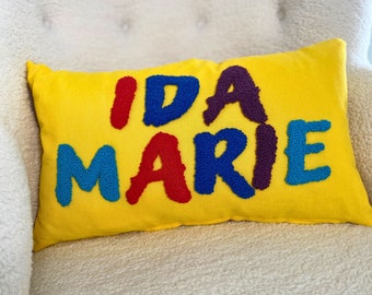 May include: A yellow rectangular pillow with the name "IDA MARIE" embroidered in red, blue, purple, and blue. The pillow is resting on a white, textured armchair.