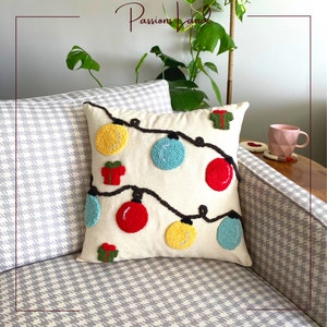 Puede incluir: Una almohada blanca con un dise&ntilde;o de luces de Navidad con adornos y regalos rojos, amarillos y azules. La almohada est&aacute; sobre un sof&aacute; de estampado gris y blanco.