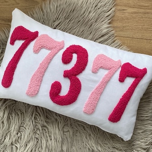 May include: A white rectangular pillow with the numbers "77377" embroidered in shades of pink. The numbers are in a textured, raised style, and the pillow is resting on a fluffy, light-colored surface.