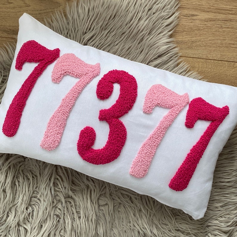 May include: A white rectangular pillow with the numbers "77377" embroidered in shades of pink. The numbers are in a textured, raised style, and the pillow is resting on a fluffy, light-colored surface.