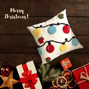 Puede incluir: Una almohada blanca con un dise&ntilde;o navide&ntilde;o festivo. La almohada presenta una cadena de luces con adornos rojos, amarillos y azules, y una peque&ntilde;a caja de regalo. La almohada est&aacute; sobre una superficie de madera con otras decoraciones navide&ntilde;as.