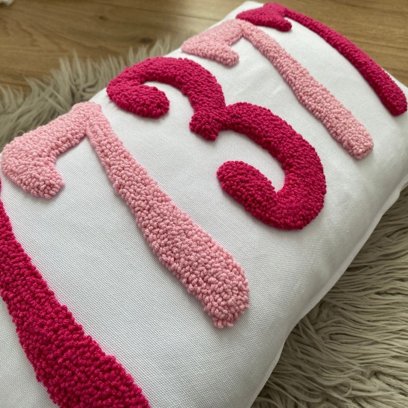 May include: White rectangular pillow with the number "137" embroidered in varying shades of pink. The numbers are raised and textured, creating a tactile design. The pillow is placed on a light-colored, textured surface.