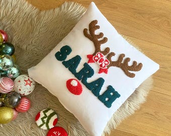 Puede incluir: Almohada blanca con un diseño de reno verde y marrón y el nombre "SARAH" en letras de lana verde. El reno tiene una nariz roja y un lazo rojo con lunares blancos.