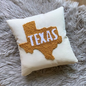 K&ouml;nnte beinhalten: Ein wei&szlig;es Kissen mit einer gelben Umrandung des Bundesstaates Texas und dem Wort "TEXAS" in wei&szlig;en Buchstaben innerhalb der Umrandung.