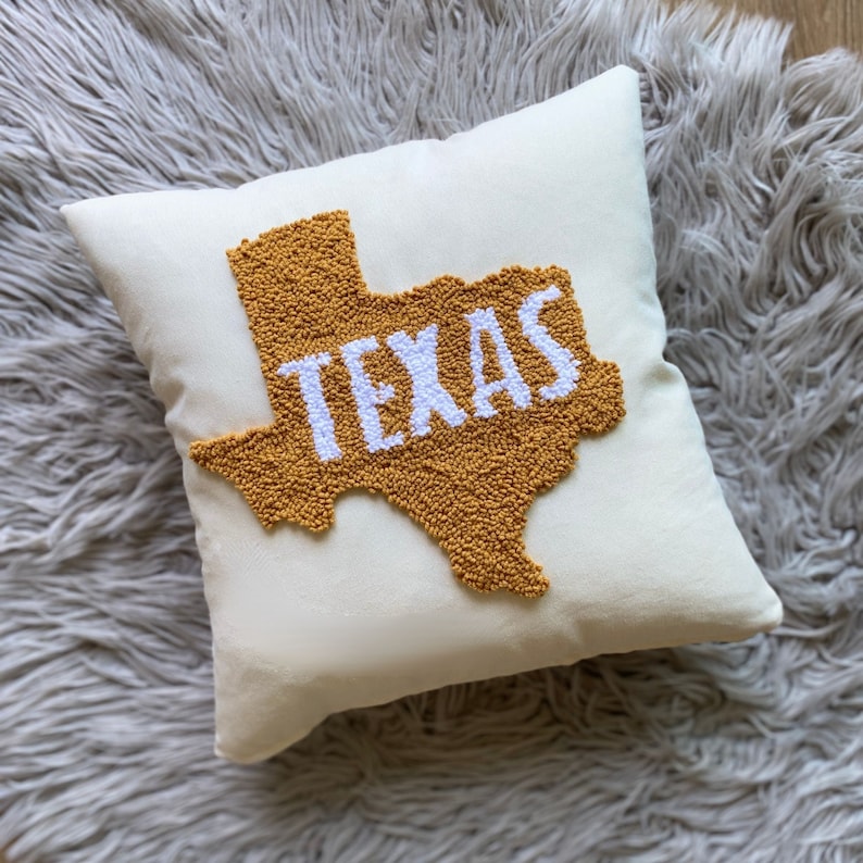 K&ouml;nnte beinhalten: Ein wei&szlig;es Kissen mit einer gelben Umrandung des Bundesstaates Texas und dem Wort "TEXAS" in wei&szlig;en Buchstaben innerhalb der Umrandung.