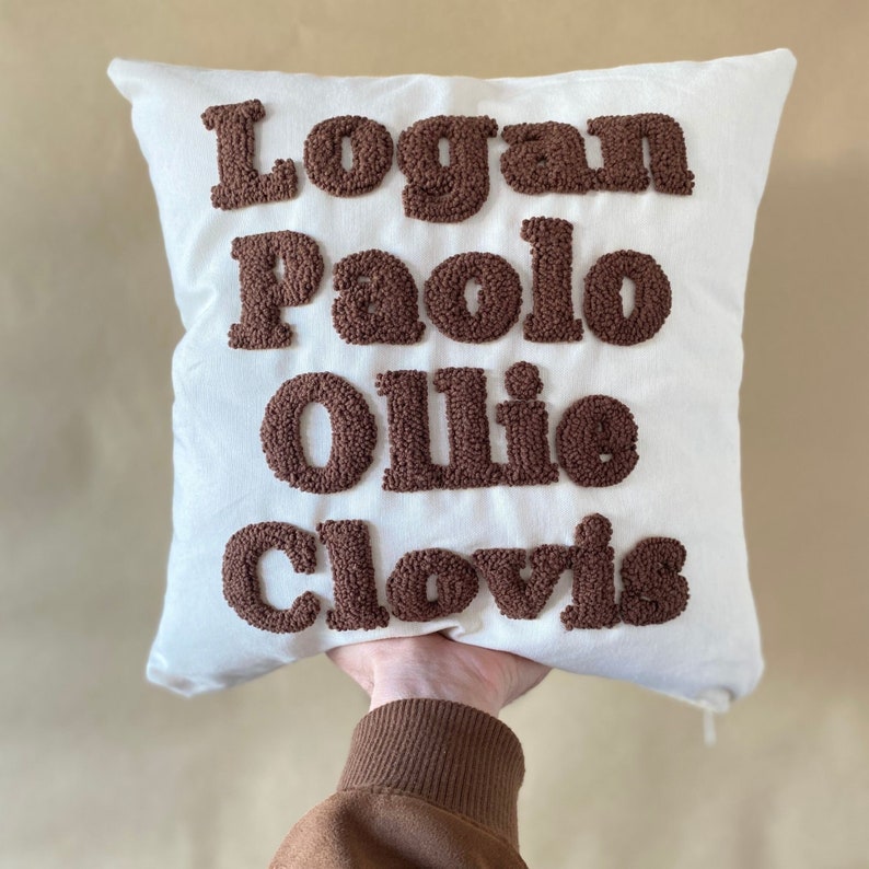 May include: White pillow with brown embroidered lettering that reads "Logan Paolo Ollie Clovis".