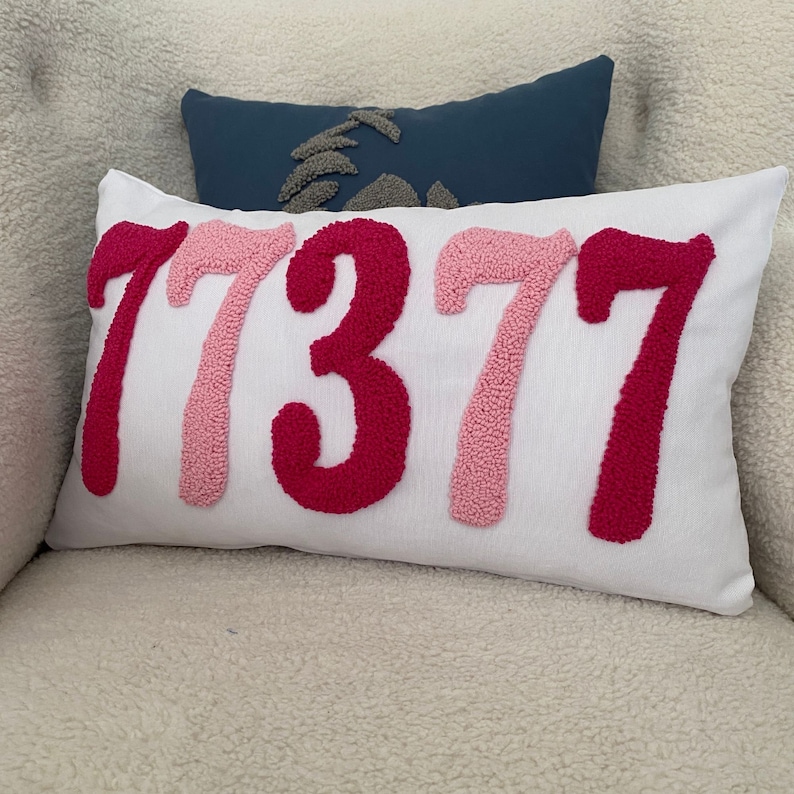 May include: White rectangular pillow with the number "77377" embroidered in shades of pink and red. A smaller blue pillow with a gray design is behind it. The pillows are on a white, textured surface.