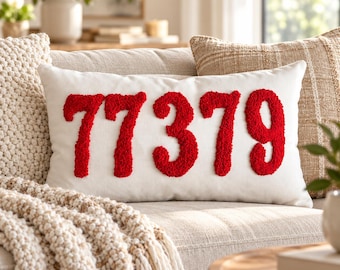 May include: A rectangular white throw pillow featuring the number 77379 in large, raised, red textured embroidery. The numbers have a fuzzy, tufted appearance and are centered on the front of the pillow, which is made of a smooth fabric.