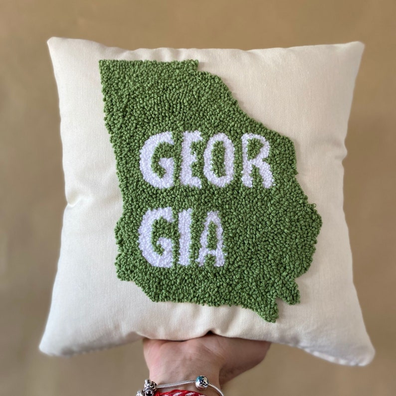 K&ouml;nnte beinhalten: Ein wei&szlig;es Kissen mit einer gr&uuml;nen Umrandung des Bundesstaates Georgia und dem Wort "GEORGIA" in wei&szlig;en Buchstaben.