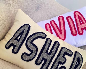 May include: Two white throw pillows with the names "Asher" and "Ivia" embroidered in colorful yarn. The letters are puffy and raised, giving the pillows a unique and textured look.