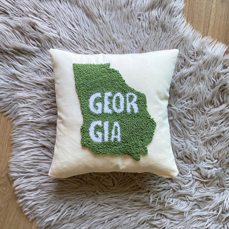 K&ouml;nnte beinhalten: Ein cremefarbenes Kissen mit einer gr&uuml;nen Umrandung des Bundesstaates Georgia und dem Wort "GEORGIA" in wei&szlig;en Buchstaben.