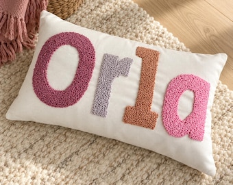 May include: A rectangular white throw pillow featuring the name Orla in large, textured, punch needle letters. The letter O is dark pink, the letter r is light purple, the letter l is terracotta orange, and the letter a is light pink.