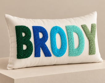 May include: A rectangular off-white throw pillow featuring the name BRODY in raised, textured punch needle embroidery. The letters are colored in a gradient from dark forest green to royal blue, bright cyan, teal, and light olive green.