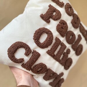 May include: A white pillow with the words "Coffee Clouds" embroidered in brown yarn. The letters are made of fluffy, looped yarn.