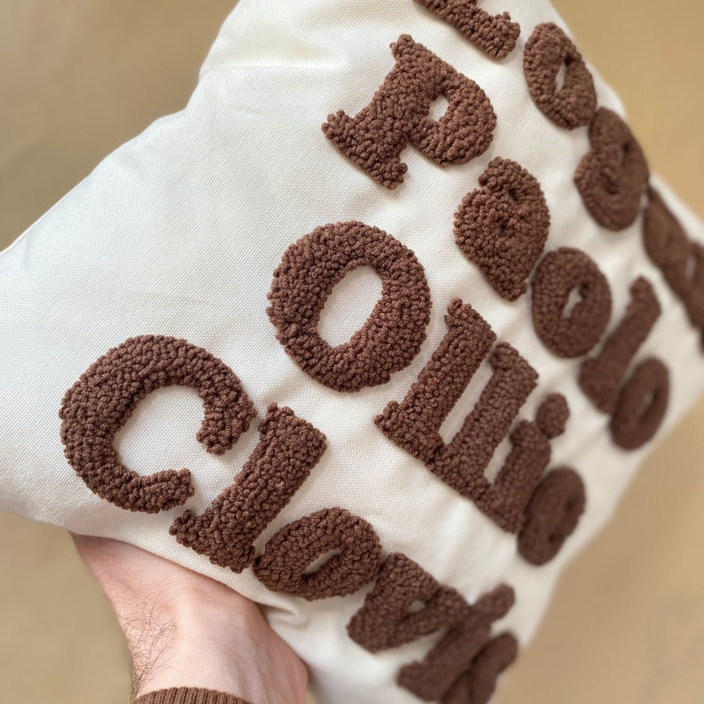 May include: A white pillow with the words "Coffee Clouds" embroidered in brown yarn. The letters are made of fluffy, looped yarn.
