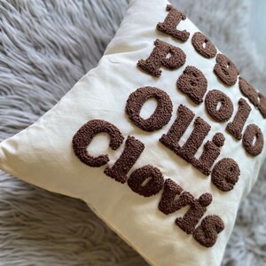 May include: White pillow with brown embroidered letters that spell "I love Paolo Ollie Clovis".