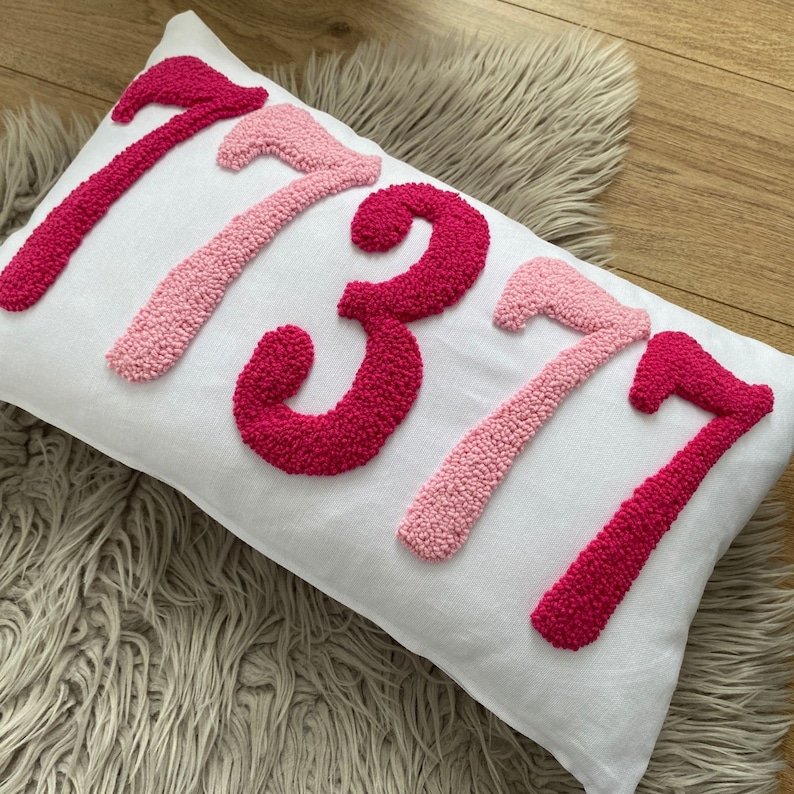 May include: White rectangular pillow with the number "7377" embroidered in shades of pink. The numbers are in a textured, raised design. The pillow is on a light-colored, textured surface.