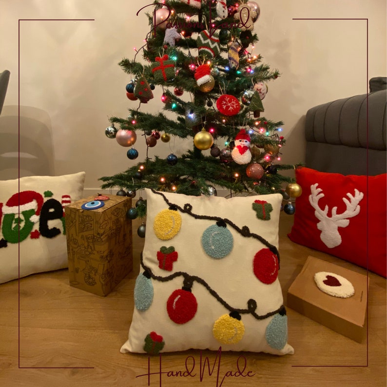Puede incluir: Una almohada blanca con un dise&ntilde;o de luces de Navidad con adornos rojos, amarillos y azules. La almohada est&aacute; hecha a mano y tiene la palabra "Hand Made" escrita en la parte inferior. La almohada est&aacute; sentada en un suelo de madera frente a un &aacute;rbol de Navidad.