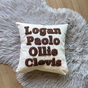 May include: A white pillow with the names "Logan", "Paolo", "Ollie", and "Clovis" embroidered in brown puffy letters.