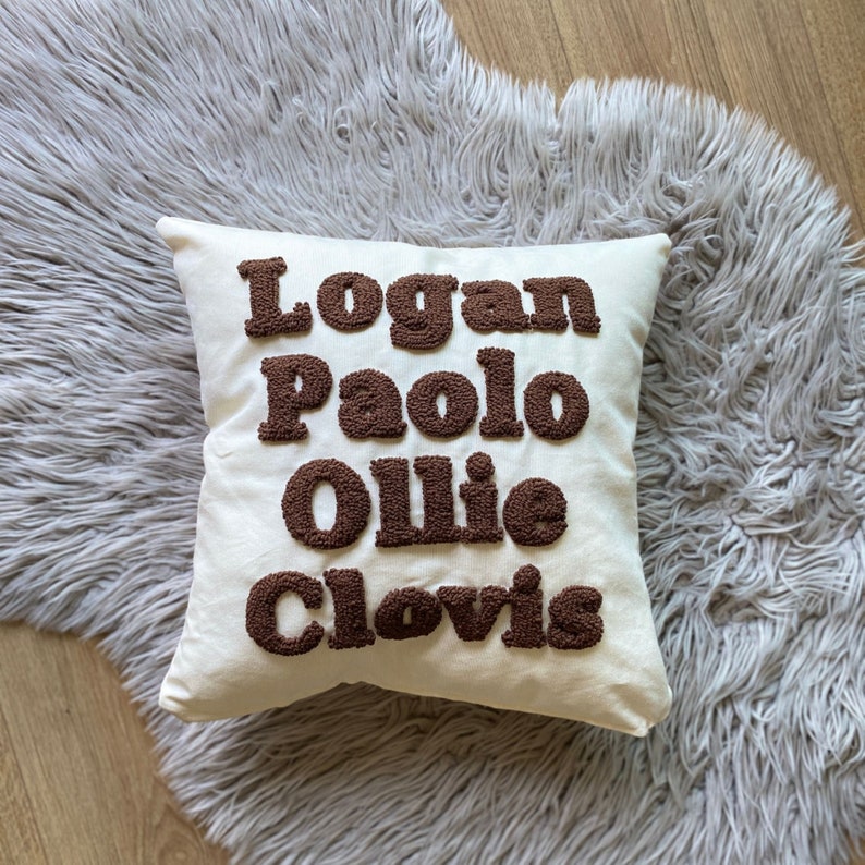 May include: A white pillow with the names "Logan", "Paolo", "Ollie", and "Clovis" embroidered in brown puffy letters.