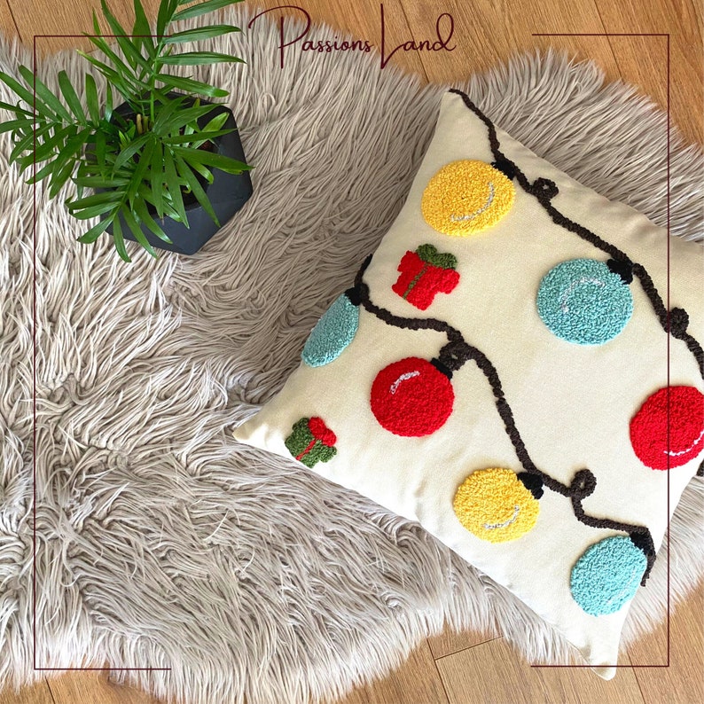 Puede incluir: Una almohada blanca con una cadena de luces navide&ntilde;as marrones y adornos coloridos. Los adornos son rojos, amarillos, azules y verdes. La almohada est&aacute; sobre una alfombra gris esponjosa.