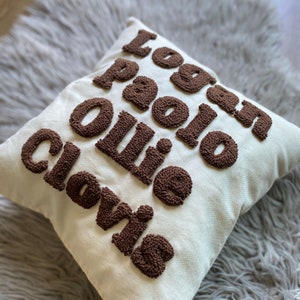 May include: A white pillow with the names Logan, Paolo, Ollie, and Clovis embroidered in brown yarn.
