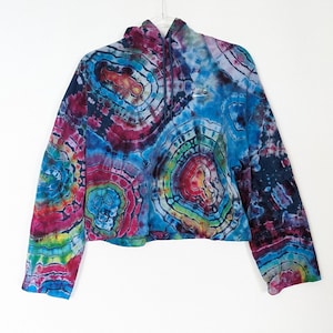 Pode incluir: Um moletom cropped tie-dye em tons de azul, roxo, vermelho e verde pendurado em um cabide de metal. O moletom com capuz apresenta um capuz com cordão e o logotipo da Nike branco no peito. As mangas compridas têm uma bainha com faixa.