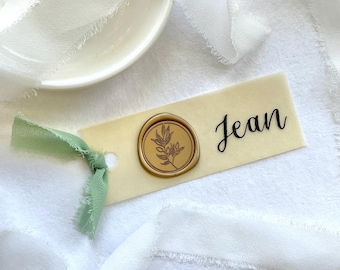 Può includere: Un cartellino rettangolare in carta color crema con il nome Jean in corsivo nero, un sigillo in ceralacca dorato circolare con un motivo a foglie botaniche e un nastro sfilacciato verde salvia annodato sul lato sinistro.