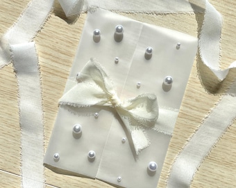 May include: Ivory-colored envelope adorned with scattered faux pearls and a matching ribbon bow. The envelope is wrapped with a frayed-edge ribbon, creating a delicate and elegant aesthetic. The pearls vary in size, adding visual interest.