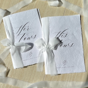 May include: Two white vow books tied with white ribbon. The text "Her Vows" and "His Vows" are written in elegant calligraphy. Each book is personalized with the couple's initials and wedding date.