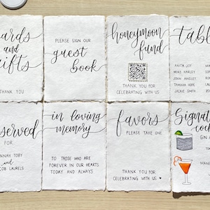 Peut inclure: Ensemble de huit panneaux en papier faits à la main avec calligraphie noire. Les panneaux comprennent des phrases telles que « Cartes et cadeaux », « Livre d'or » et « Fonds de lune de miel ». Parfait pour un mariage.