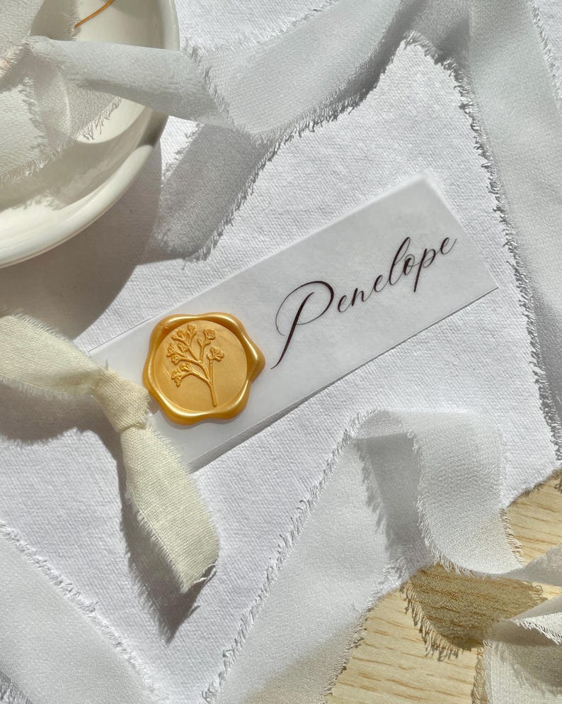 Pu&ograve; includere: Un segnaposto rettangolare bianco con il nome Penelope scritto in un'elegante calligrafia nera. Un sigillo di ceralacca dorato con un motivo floreale botanico &egrave; fissato sul lato sinistro, con un nastro di seta bianco sfrangiato annodato.