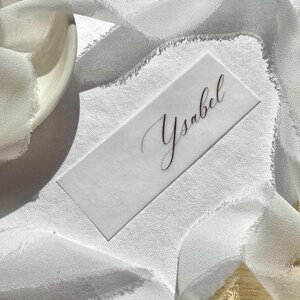 Pu&ograve; includere: Un segnaposto rettangolare bianco con il nome Ysabel scritto in un'elegante calligrafia scura. Il cartoncino &egrave; posizionato su una superficie bianca testurizzata e parzialmente avvolto da un lungo nastro bianco fluido con bordi sfilacciati.