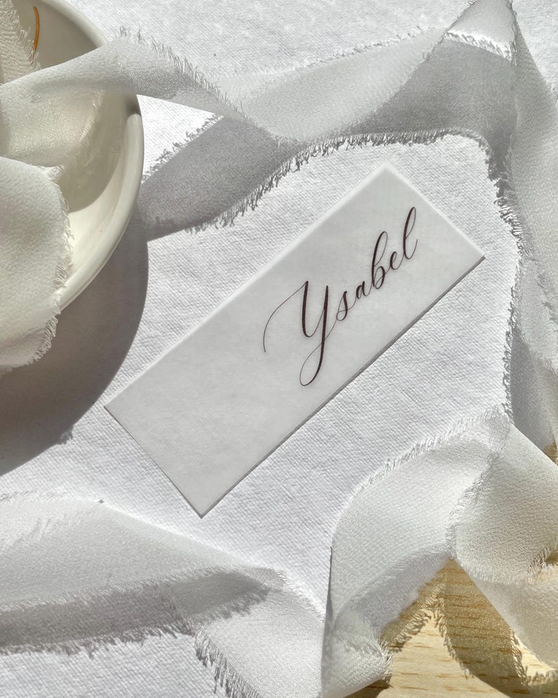 Pu&ograve; includere: Un segnaposto rettangolare bianco con il nome Ysabel scritto in un'elegante calligrafia scura. Il cartoncino &egrave; posizionato su una superficie bianca testurizzata e parzialmente avvolto da un lungo nastro bianco fluido con bordi sfilacciati.