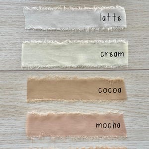 Pu&ograve; includere: Sei strisce di nastro in tessuto con bordi sfilacciati disposte orizzontalmente. I colori sono etichettati come white, latte, cream, cocoa, mocha e chestnut. Ogni striscia presenta una consistenza opaca morbida e un bordo grezzo distintivo.