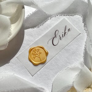 Pu&ograve; includere: Un segnaposto rettangolare bianco con il nome Erika in elegante corsivo nero, decorato con un sigillo di ceralacca dorato circolare con un delicato motivo floreale in rilievo, circondato da un nastro di seta bianco con bordi sfrangiati.