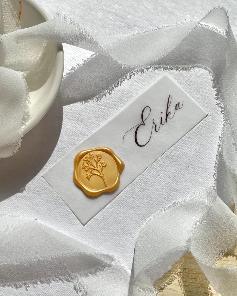 Pu&ograve; includere: Un segnaposto rettangolare bianco con il nome Erika in elegante corsivo nero, decorato con un sigillo di ceralacca dorato circolare con un delicato motivo floreale in rilievo, circondato da un nastro di seta bianco con bordi sfrangiati.