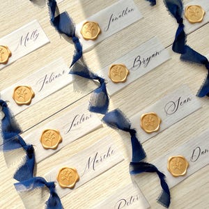 Pu&ograve; includere: Una collezione di segnaposto rettangolari in carta bianca con nomi scritti a mano in inchiostro nero, ciascuno decorato con un sigillo di ceralacca dorato con motivo floreale e legato con un nastro di seta blu scuro sfilacciato, disposti.