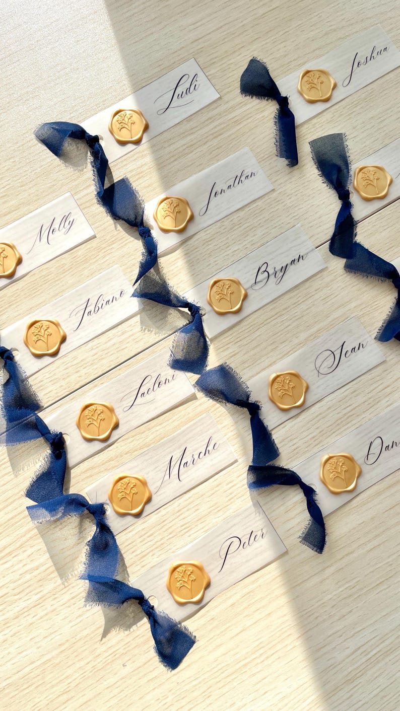 Pu&ograve; includere: Una collezione di segnaposto rettangolari in carta bianca con nomi scritti a mano in inchiostro nero, ciascuno decorato con un sigillo di ceralacca dorato con motivo floreale e legato con un nastro di seta blu scuro sfilacciato, disposti.