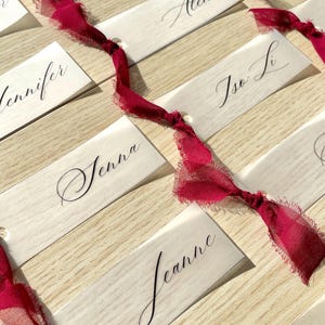 Pu&ograve; includere: Una collezione di segnaposto rettangolari in carta bianca con nomi in calligrafia nera, tra cui Araceli, Scout, Ryan, Lucia, Alene, Jennifer, Tse Li, Jenna e Jeanne. Ogni carta &egrave; legata con un nastro di seta rosso scuro sfilacciato.