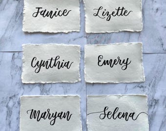 May include: Six rectangular off-white paper cards with deckled edges, each featuring a name written in black calligraphy. The names displayed are Janice, Lizette, Cynthia, Emery, Maryan, and Selena, arranged in two columns of three cards each.