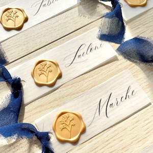 Pu&ograve; includere: Segnaposto rettangolari bianchi con nomi scritti a mano come Molly, Fabiano, Leoni e Marche. Ogni carta &egrave; decorata con un sigillo di ceralacca dorata con un motivo floreale e un nastro di seta blu scuro sfilacciato e fluente.