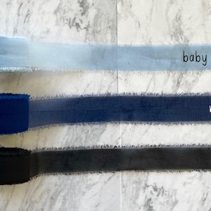 Pu&ograve; includere: Tre rocchetti di nastro con bordi sfilacciati disposti orizzontalmente. Il nastro superiore &egrave; azzurro, quello centrale &egrave; blu navy e quello inferiore &egrave; nero. Ogni rocchetto &egrave; etichettato con il nome del colore in semplice testo nero.