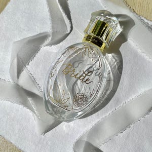 Puede incluir: Frasco de perfume ovalado de cristal transparente con detalles dorados y tapón de cristal. La botella presenta la palabra "Bride" en escritura dorada, con diseños florales. Sobre un fondo de tela blanca.