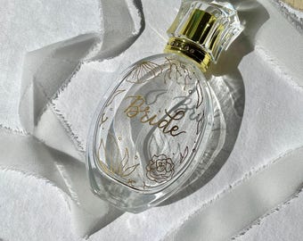 Può includere: Flacone di profumo ovale in vetro trasparente con dettagli dorati e tappo di cristallo. La bottiglia presenta la scritta "Bride" in oro, con disegni floreali. Posizionata su uno sfondo di tessuto bianco.