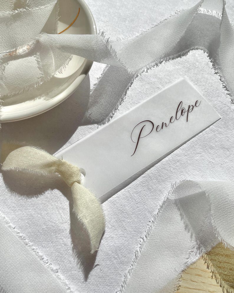 Pu&ograve; includere: Un cartoncino rettangolare bianco con il nome Penelope stampato in un carattere corsivo scuro. Il cartoncino presenta un foro sul lato sinistro con un nastro in tessuto sfilacciato color crema annodato, appoggiato su una superficie in tessuto bianco.