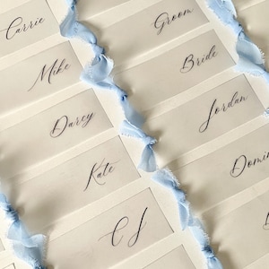 Pu&ograve; includere: Una collezione di segnaposto rettangolari bianchi con nomi in calligrafia nera come Dave, Jara, Jackie, Carrie, Groom, Mike, Bride, Darcy, Jordan, Kate e Amy. Ogni carta &egrave; decorata con una striscia verticale di nastro azzurro sfilacciato.
